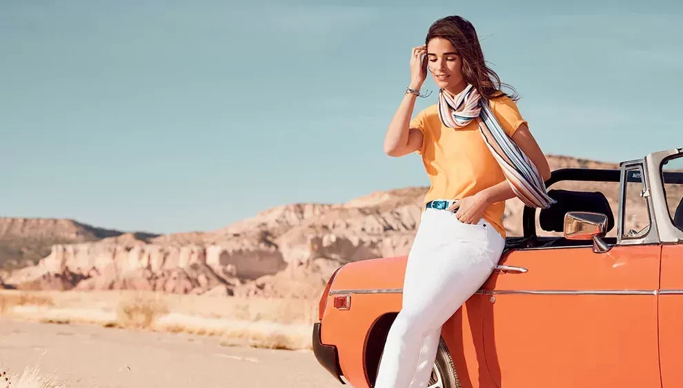 A fashion model leans against an orange car, wearing an orange top and white skinny jeans